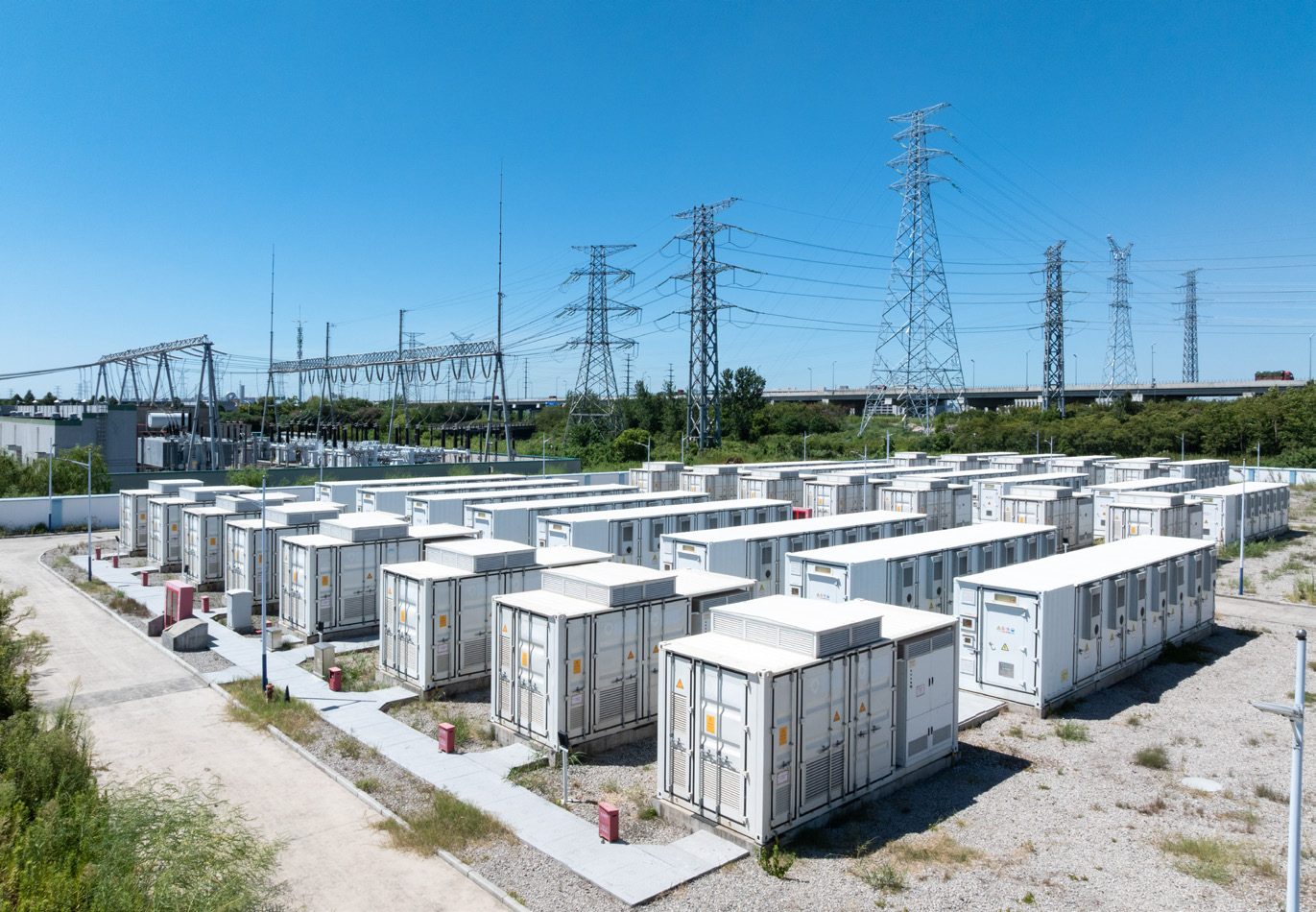 Große stationäre Batteriespeicher auf einem Betriebsgelände mit Strommasten und Umspanntechnik im Hintergrund.