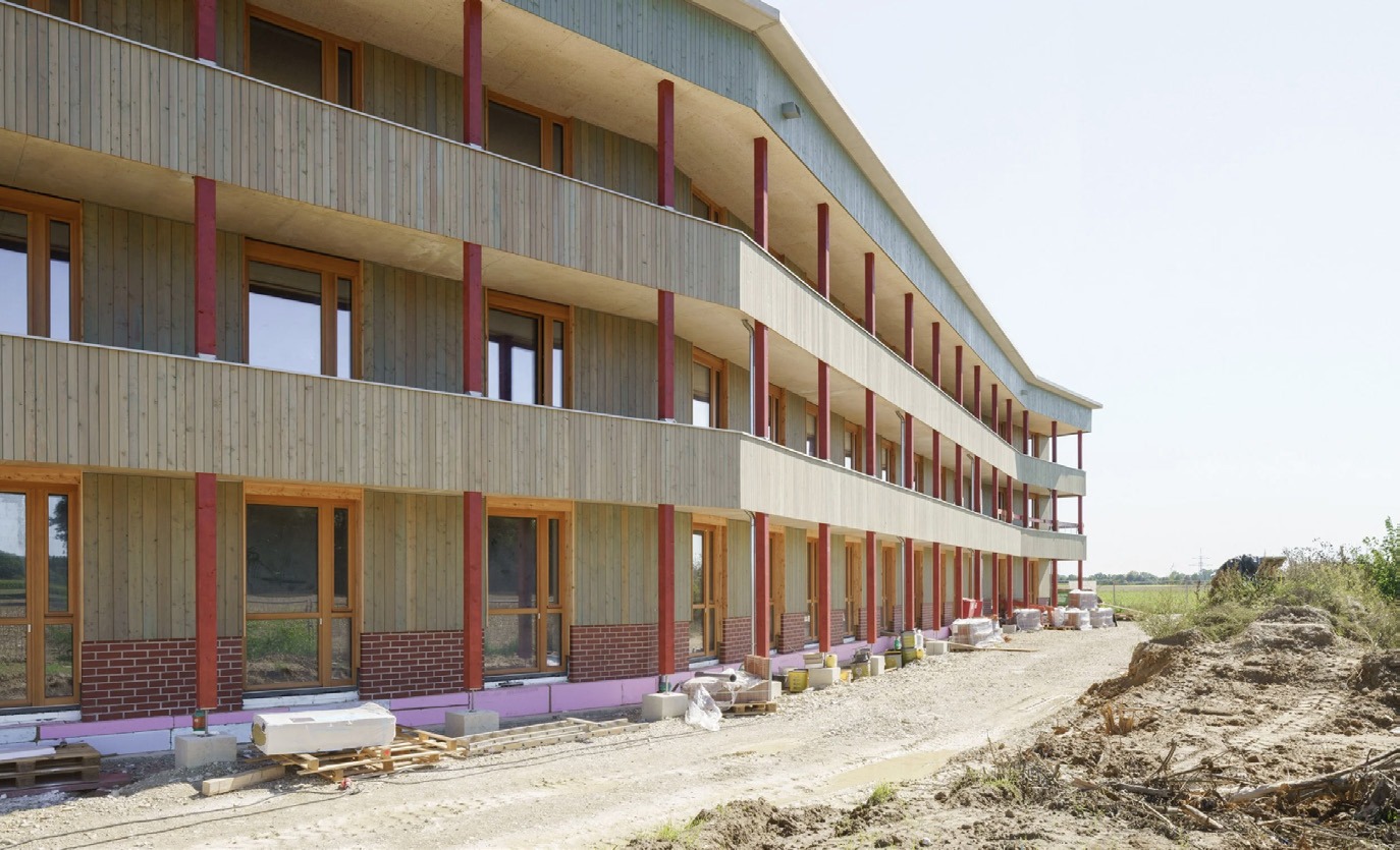 Mehrgeschossiges Wohngebäude in Holzbauweise während der Bauphase, mit umlaufenden Balkonen und roten Stützen.