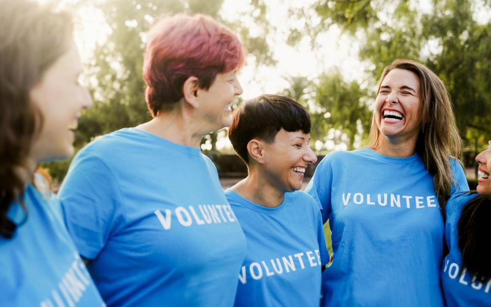 Menschengruppe mit t shirt mit der Aufschrift Volunteers stehen eingehakt nebeinander und lachen 